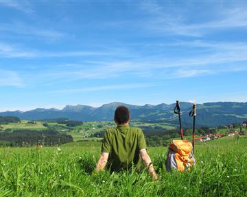 A hiker sits on a green meadow and looks at the mountains. The sky is clear and blue, in the background there are gentle hills and a small village visible.