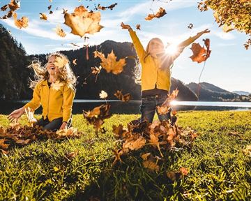 Zwei Kinder in gelben Jackets spielen fröhlich im Herbstlaub. Sie werfen bunte Blätter in die Luft, während die Sonne im Hintergrund scheint.