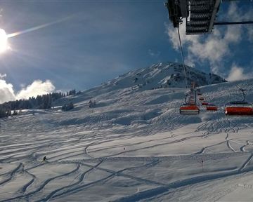 Ein schneebedeckter Hang mit Skizügen und einer sonnigen, klaren Himmel. Im Hintergrund ist ein Skilift zu sehen.