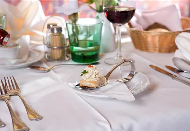 An elegant table with cutlery, a glass of wine, and a small portion on a spoon. In the background, flowers and soft colors are visible.