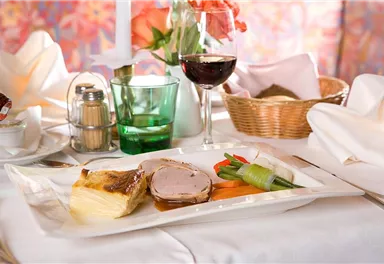 An elegant dish with meat, vegetables, and potatoes on a white plate. Accompanied by a glass of red wine and decorated table elements.