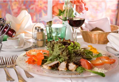 A freshly prepared dish with chicken fillet, colorful salad, and slices of tomato. In the background, a glass of red wine and a decorative table setting can be seen.