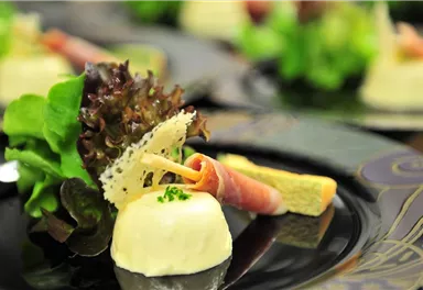 A stylish dish with a creamy main ingredient, surrounded by fresh salad and ham. The arrangement on an elegant black plate ensures an appealing presentation.