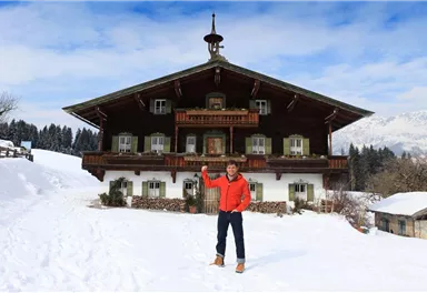 Ein schönes Holzhaus in den Bergen, umgeben von Schnee und Bäumen. Eine Person steht davor und hebt die Hand zur Begrüßung.