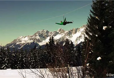 A visitor glides on a zip line over a snow-covered landscape. In the background, majestic mountains and fir trees can be seen.