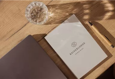 Ein minimalistisches Arbeitsumfeld mit einem offenen Notizbuch und einem Laptop. Nebenbei steht ein Glas Wasser auf einem Holztisch.