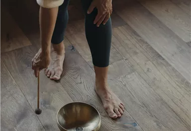 Eine Person steht barfuß auf einem Holzboden und spielt mit einem Stock auf zwei Klangschalen. Die Atmosphäre wirkt entspannt und meditative.