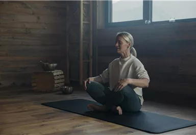 Eine Frau sitzt meditierend auf einer Yogamatte in einem ruhigen, holzverkleideten Raum. Sanftes Licht strahlt durch die Fenster, während sie entspannt aussieht.