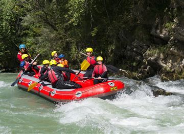 Eine Gruppe von Menschen fährt in einem roten Raft über einen wilden Fluss. Alle tragen Helme und Schwimmwesten und scheinen viel Spaß zu haben.