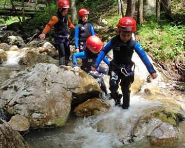 Vier Kinder mit Helmen und Neoprenanzügen waten durch einen kleinen Bach. Um sie herum sind Felsen und üppige grüne Bäume.