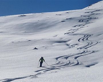 Eine verschneite Berglandschaft mit einem Skifahrer, der frische Spuren im Schnee hinterlässt. Im Hintergrund sind schneebedeckte Gipfel und ein blauer Himmel zu sehen.