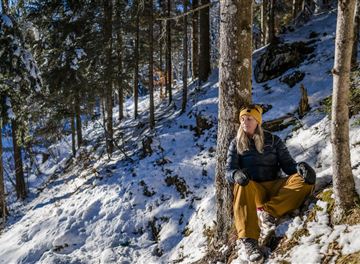 Eine Person sitzt entspannt in einem verschneiten Wald. Um sie herum stehen hohe Bäume und die Sonne scheint durch die Äste.