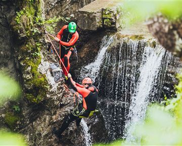 Zwei Personen in Neoprenanzügen klettern an einer felsigen Wand neben einem Wasserfall. Sie nutzen Seile zur Sicherung und arbeiten eng zusammen.