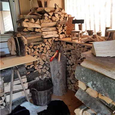 A cozy room with stacked firewood and a wooden log. A wooden chair and a basket complement the rustic atmosphere.