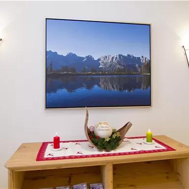 A cozy room with a wooden shelf and a landscape painting above a still body of water. On the shelf are candles and a decorative bowl.