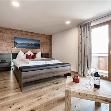 A modern bedroom with wooden paneling and large windows. There is a large bed, a cozy seating area, and a table with a wine bottle and glasses.