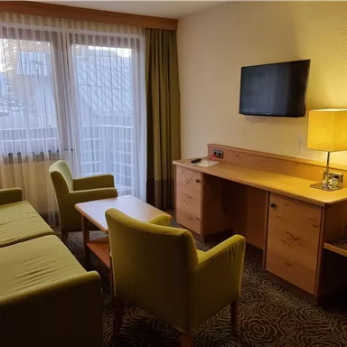 Ein gemütliches Hotelzimmer mit zwei grünen Stühlen, einer Couch und einem Tisch. Es gibt einen Schreibtisch und einen Fernseher an der Wand, sowie große Fenster, die viel Licht hereinlassen.