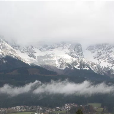 A majestic mountain landscape with snow-capped peaks and a cloudy sky. Below lie green meadows and a small village.