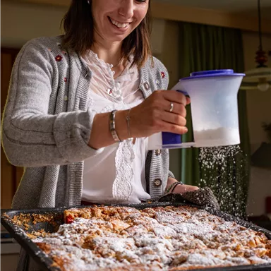 Eine lächelnde Frau bestreut einen frisch gebackenen Kuchen mit Puderzucker. Der Kuchen liegt auf einem Backblech in einer gemütlichen Küche.
