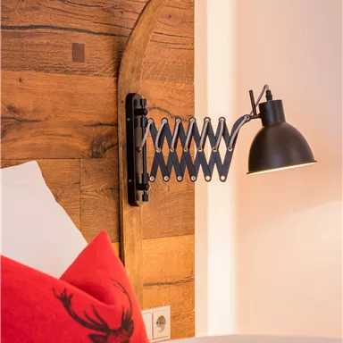 An elegant wall lamp is attached to a wooden panel adorned with a slate edge. In the foreground lies a raw cushion with a decorative design.