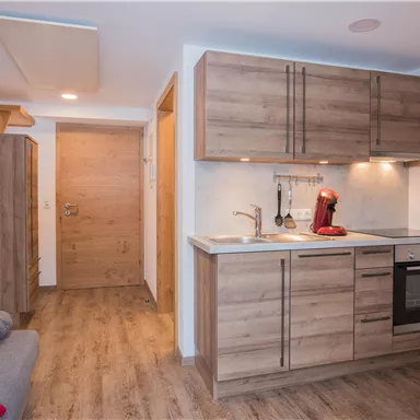 A modern kitchen with wooden cabinets and a stove. In the background, the entrance is visible.