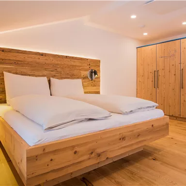 A modern bedroom with a cozy wooden bed and large pillows. The room has a warm, inviting wooden floor and a bright, friendly atmosphere.