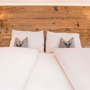 A cozy double bed with white bedspreads and decorative pillows. In the background, there is wooden paneling with warm light sources.