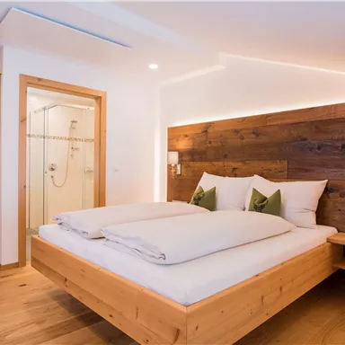A modern bedroom with a wooden double bed and stylish cushions. In the background, a bathroom with a glass shower cabin can be seen.