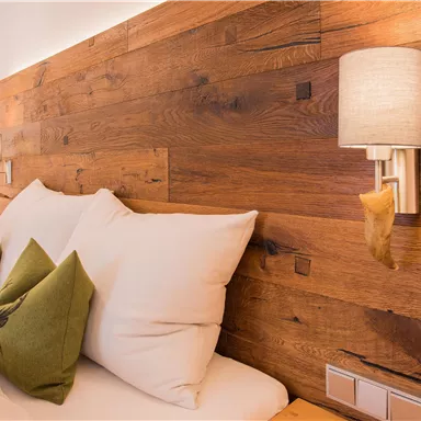 A cozy bedroom with wood paneling and a soft bed. On the pillows are green decorative cushions, and the walls are illuminated with warm light.