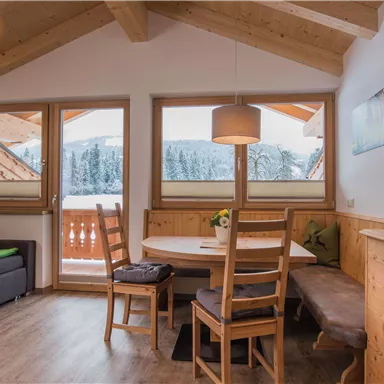 A cozy living space with wooden furniture and large windows. Outside is a snow-covered view of the winter landscape.