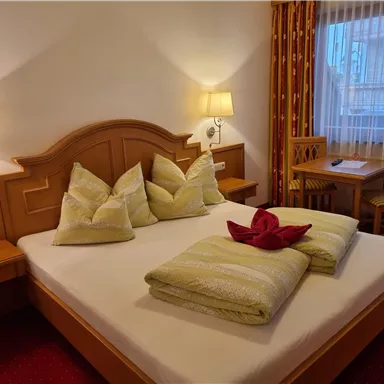 Ein gemütliches Schlafzimmer mit einem großen Bett, auf dem bunte Kissen liegen. Neben dem Bett steht ein Holztisch und Stühle, und das Fenster lässt viel Licht herein.