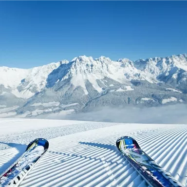 A wintry mountain landscape with snow-covered peaks and clear blue sky. In the foreground, skis lie on freshly groomed slopes.