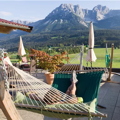 Eine Terrasse mit einer Hängematte und Blick auf die Berge. Die Umgebung ist grün und einladend, ideal zum Entspannen.