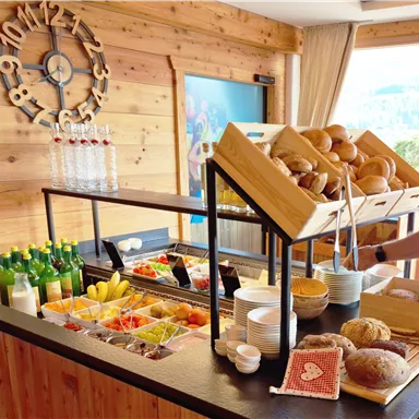 Ein Buffet mit frischen Brötchen, Obst und verschiedenen Getränken. Die Holzverkleidung und der große Fensterbereich schaffen eine einladende Atmosphäre.