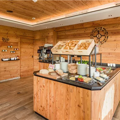 Ein einladender Frühstücksbereich mit Holzverkleidung und einem Buffet voller frischer Speisen. Auf dem Tisch sind verschiedene Brote, Aufschnitt und Obst zu sehen.