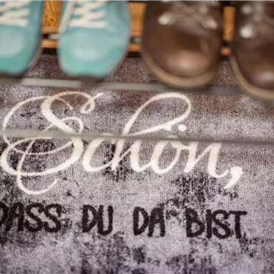 Ein Willkommensmatte mit der Aufschrift „Schön, dass du da bist“. Darüber sind ein paar Schuhe in verschiedenen Farben zu sehen.