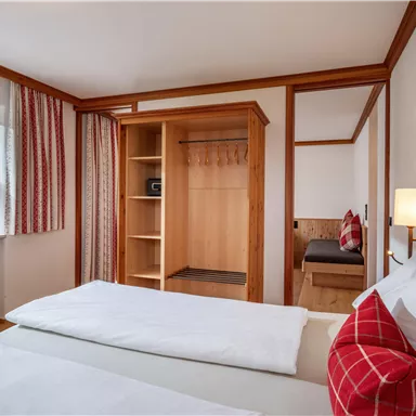 Ein gemütliches Zimmer mit zwei Betten und einem Holzschrank. Die Fenster lassen viel Tageslicht herein und die Einrichtung ist freundlich und einladend.