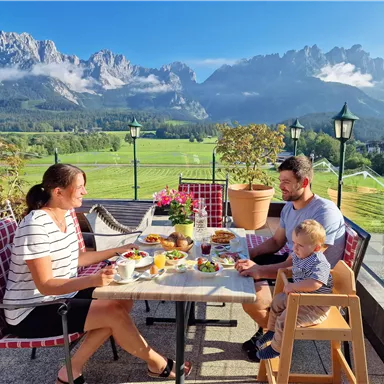 Eine Familie sitzt an einem Tisch im Freien und genießt ein Frühstück mit Blick auf die Berge. Die Landschaft ist grün und sonnig, mit blühenden Pflanzen und klarem Himmel.