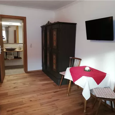Ein gemütlicher Raum mit Holzboden und hellen Wänden. Es gibt einen Tisch mit rotem Tischläufer, Stühle und einen Fernseher an der Wand.
