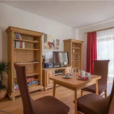 Ein gemütliches Wohnzimmer mit Holzmöbeln und einem Tisch, umgeben von Stühlen. Große Fenster mit roten Vorhängen lassen viel Licht herein.