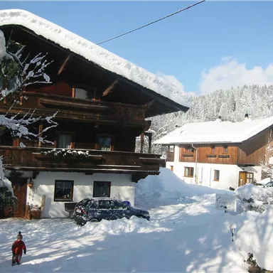 Ein malerisches altes Holzhaus im Schnee mit schneebedeckten Dächern. Im Vordergrund spielt ein Kind im tiefen Schnee.
