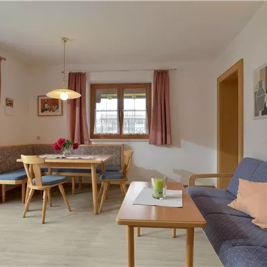 Ein gemütliches Wohnzimmer mit einer Sitzgruppe und einem Esstisch. Helle Wände und große Fenster sorgen für eine einladende Atmosphäre.