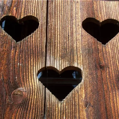 Drei herzförmige Öffnungen in einer rustikalen Holzwand. Das Licht strahlt sanft durch die Herzen.