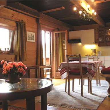 A cozy living space with wooden walls, a table, and chairs. The room has windows with curtains and a cozy atmosphere.