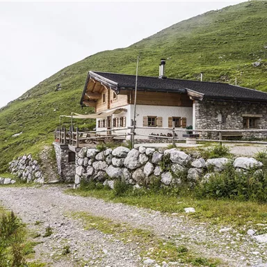 Eine gemütliche Berghütte umgeben von grünen Hügeln. Der Weg zur Hütte ist mit Kies bedeckt und lädt zum Wandern ein.