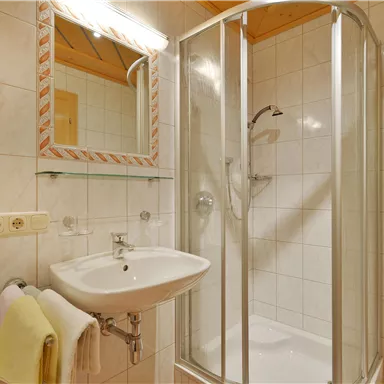 Ein modernes Badezimmer mit einer Duschkabine und einem Waschbecken. Die Wände sind mit weißen Fliesen gefliest und es gibt einen Spiegel über dem Waschbecken.