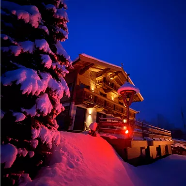 Ein gemütliches Holzhaus im Schnee, beleuchtet von warmem Licht. Rundherum sind schneebedeckte Bäume und der Himmel ist dunkelblau.