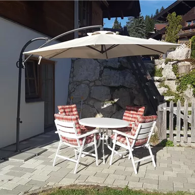 A cozy seating area with a sun umbrella and two chairs on a paved path. In the background, green plants and a wooden hedge can be seen.