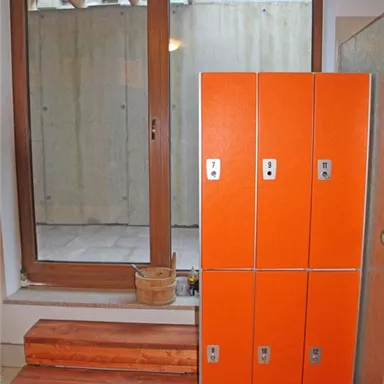 Ein Raum mit einem großen Fenster, durch das Tageslicht strömt. Rechts stehen orangefarbene Spinde und links sind Holzstufen sichtbar.