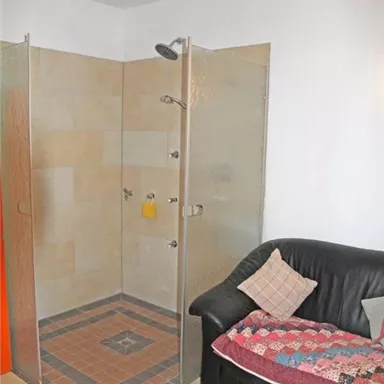 Ein Badezimmer mit einer modernen Dusche aus Glas und einem dunklen Sofa. Der Boden ist gefliest und die Wände sind hell gestrichen.